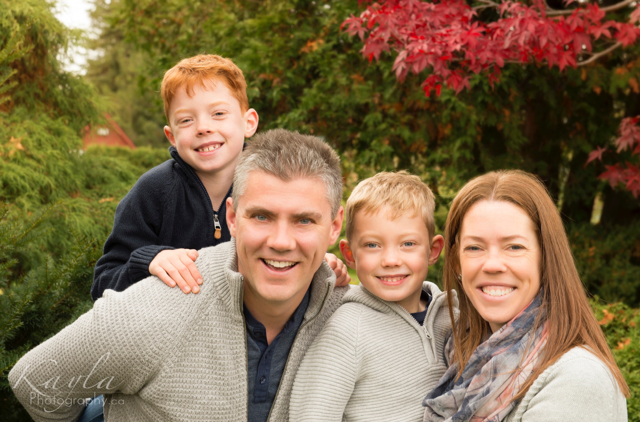 Family of four smiling together outdoors with autumn leaves, photographed by Kayla Photography in Ottawa