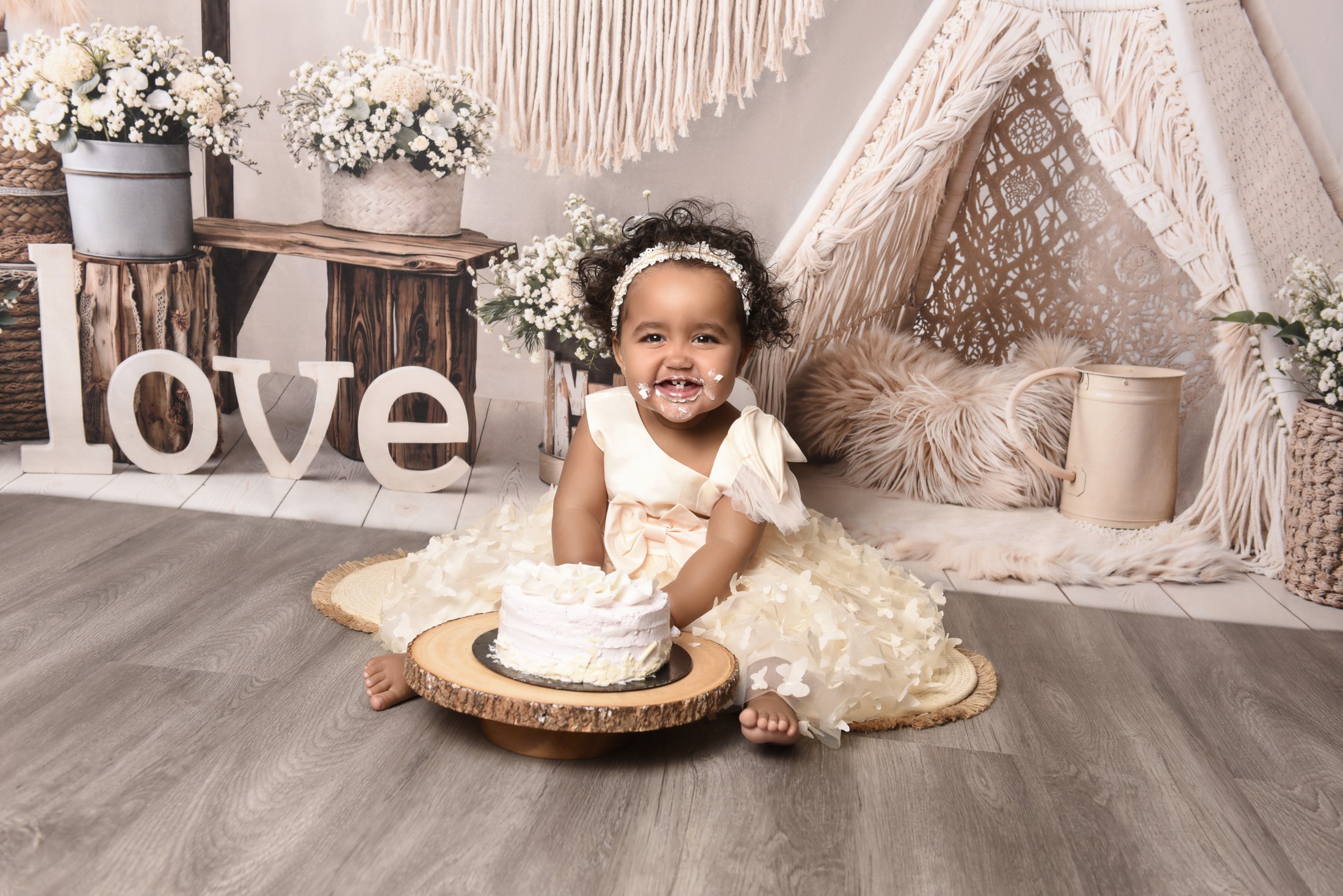 Smiling child in a cream dress enjoying a cake smash in a floral studio set